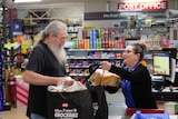 A woman behind a checkout helps a supermarket shopper load his bags.