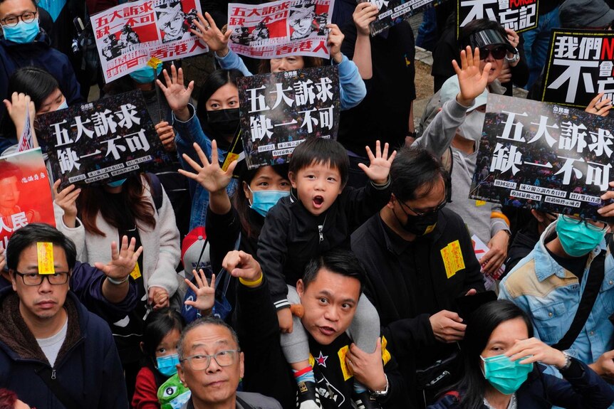 People hold up hands in the air with five fingers, including small child in the centre, holding banners with Chinese text.