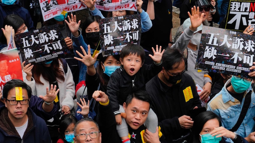 People hold up hands in the air with five fingers, including small child in the centre, holding banners with Chinese text.