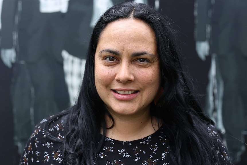 Ngarra Murray smiles in a black top in front of a mural of historic Victorian Aboriginal leaders.