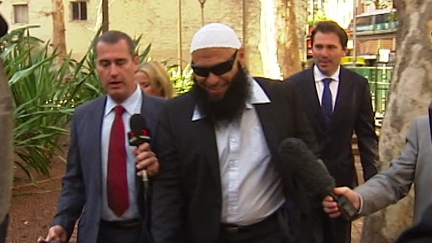 A man with a long beard wears a suit, sunglasses and an Islamic cap as he walks into a court building, surrounded by reporters.