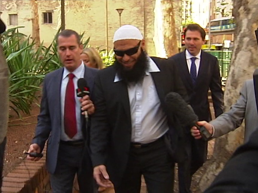 A man with a long beard wears a suit, sunglasses and an Islamic cap as he walks into a court building, surrounded by reporters.