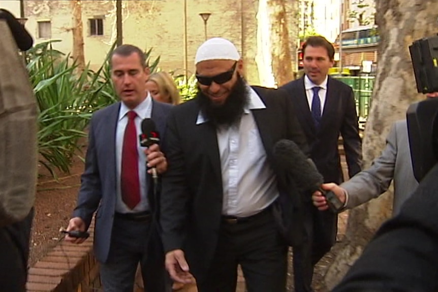 A man with a long beard wears a suit, sunglasses and an Islamic cap as he walks into a court building, surrounded by reporters.