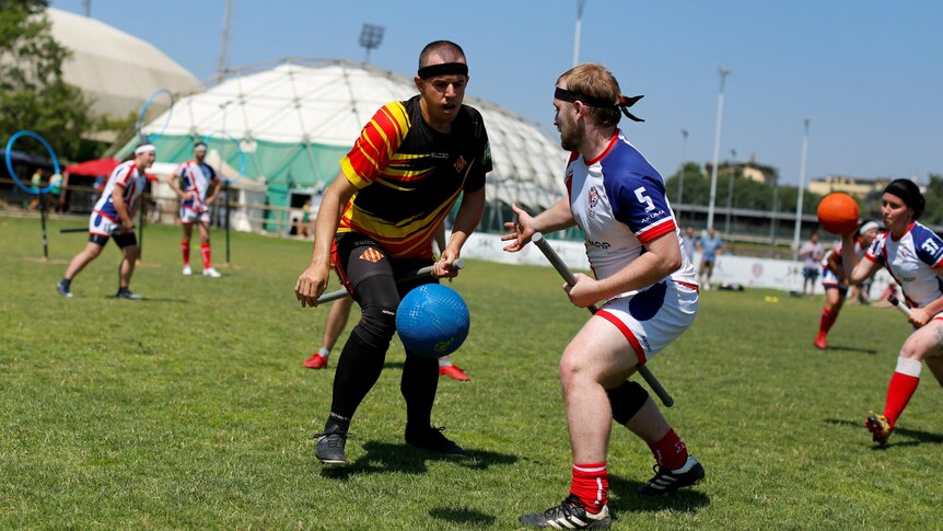 Two men are pictured in action chasing a blue ball. they are wearing sports attire and have black bands tied around their heads.