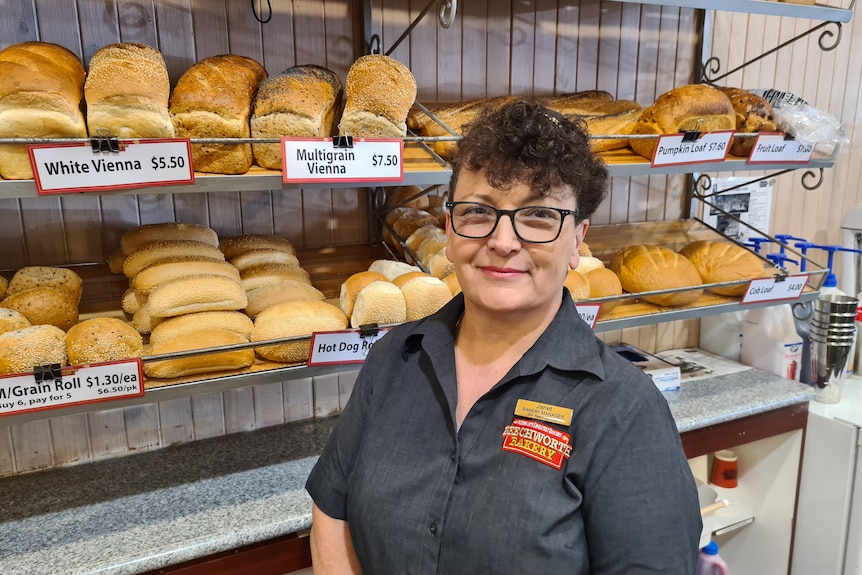 A smiling, bespectacled woman with short dark curly hair stands in a bakery.