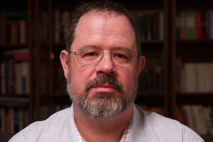 Michael Fotheringham, dressed in a shirt, stands in front of a bookcase indoors.