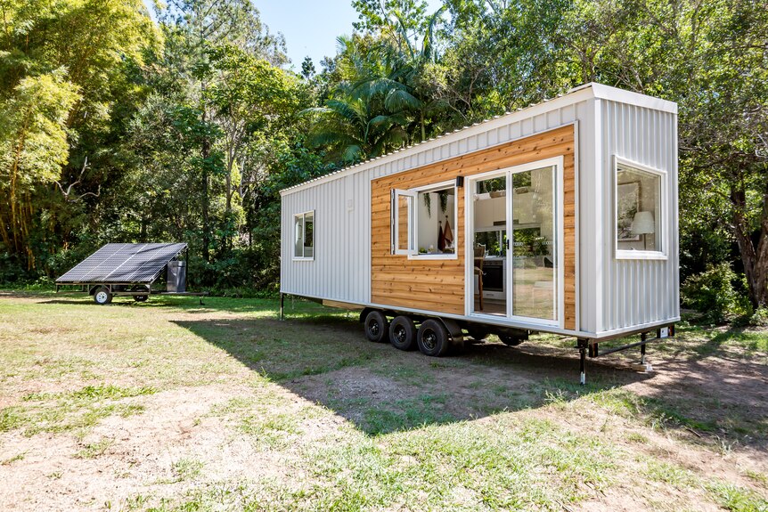 Tiny house with light brown wood and grey metal paneling parked in a clearing.