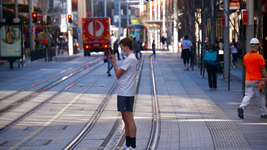 A masked man stands looking at his phone on the light rail corridor in inner Sydney.