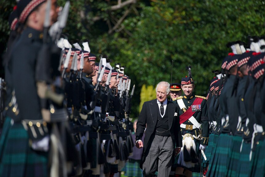 King Charles walks in the middle of a guard wearing kilts. 
