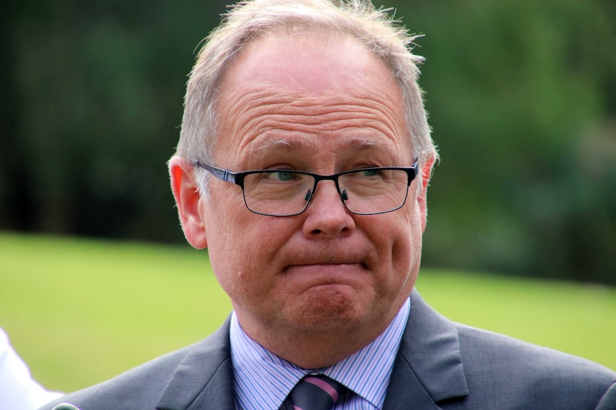 Head and shoulders image of a man in a suit, tie and glasses speaking at an outdoors press conference.