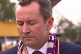 Mark McGowan with head bowed and eyes closed, wearing a suit and a Perth Glory scarf.