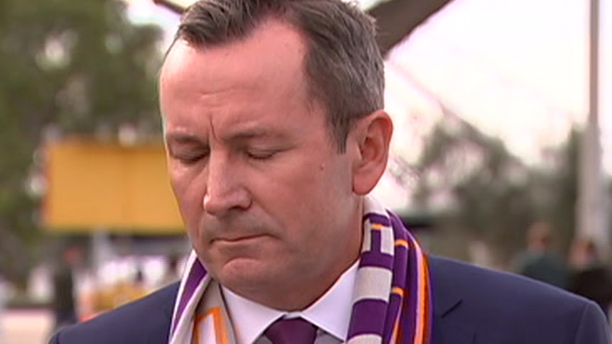 Mark McGowan with head bowed and eyes closed, wearing a suit and a Perth Glory scarf.