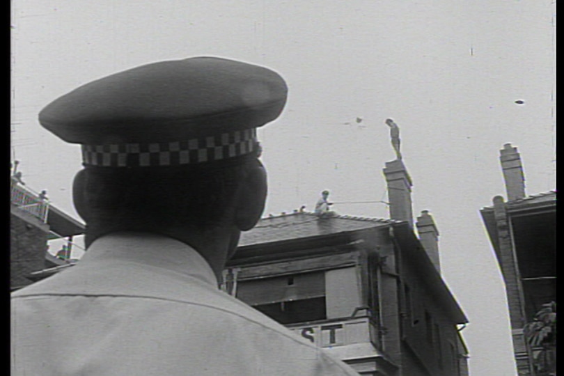 A black and white photo of the back of a police officer's head as he stares up at a man standing on a rooftop.