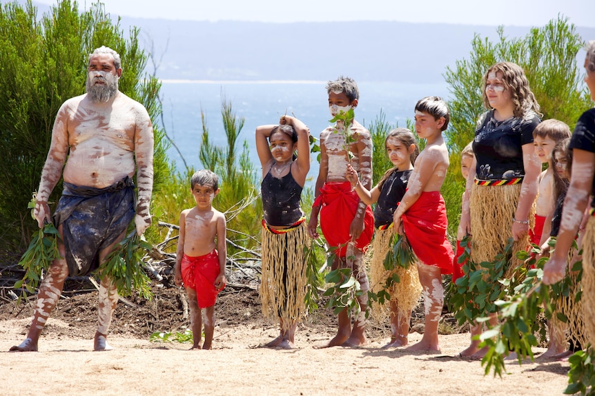 An Indigenous man in traditional dress and white body paint, with a group of children also in traditional dress.