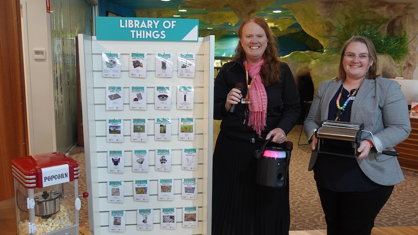 Two women pose with a karaoke machine and a pasta maker next to a board displaying items available for loan.