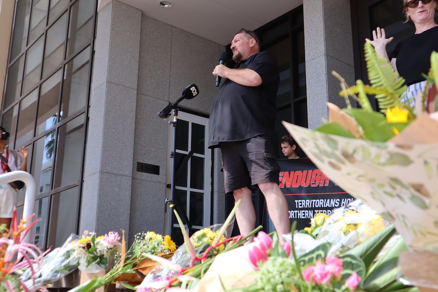 A man wearing a black t-shirt holding a microphone and speaking out the front of a marble building