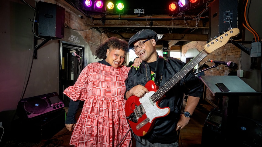 Two people, one holding an electric guitar, smile at the computer.