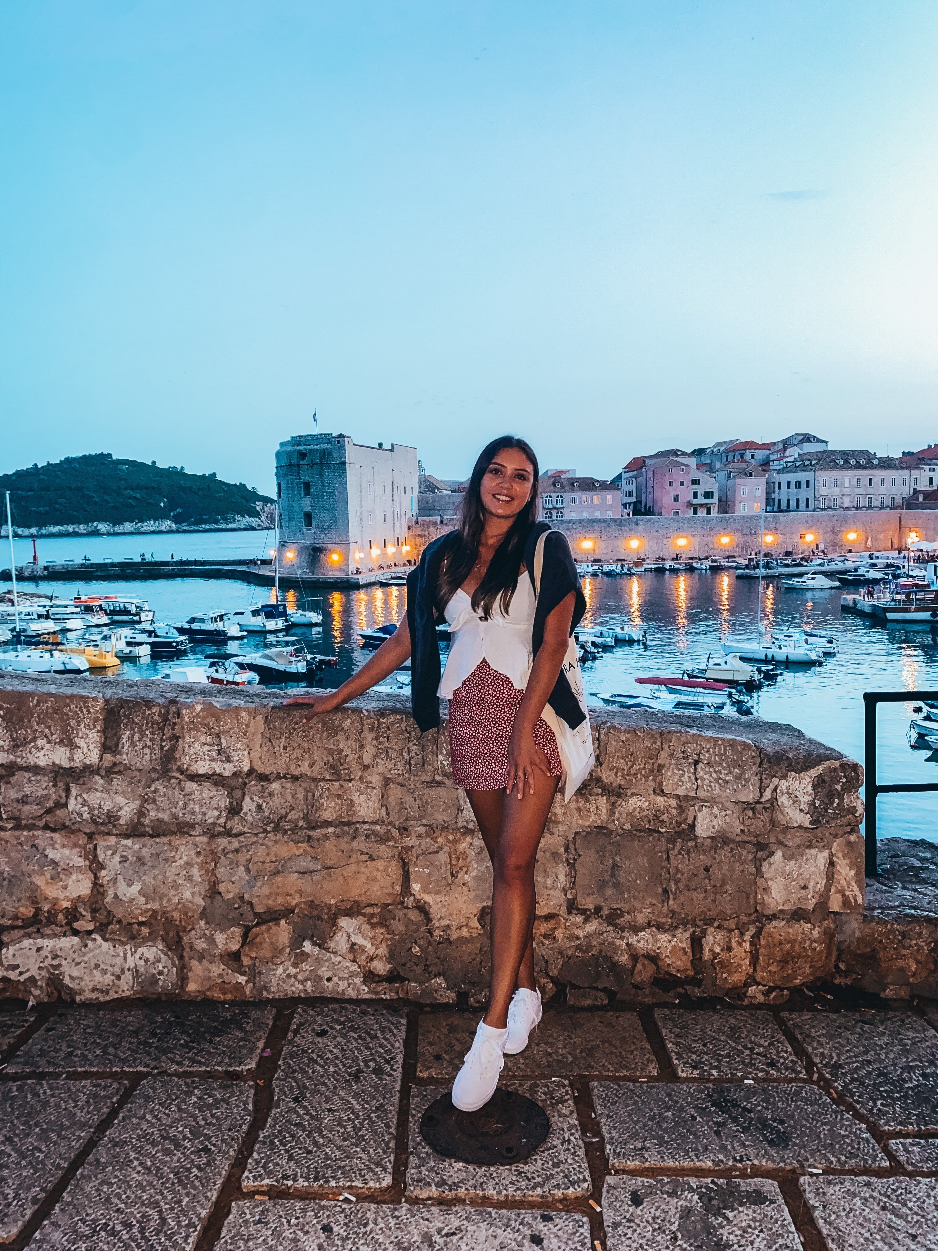 Kate smiles in front of a European harbour, wearing a red flowery skirt and a white top.