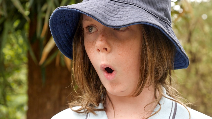 Boy with shoulder length brown hair in blue hat with lips pursed