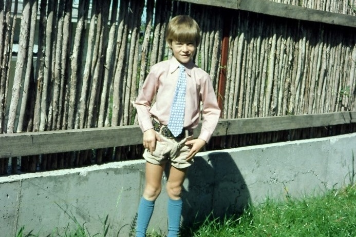 A 1970s photo of a young boy in a garden, dressed in a shirt, tie and shorts.