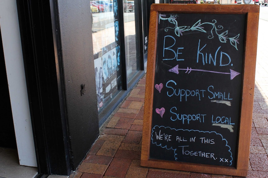 A blackboard sign on a footpath outside a coffee shop. It says be kind, support small, support local, we're all in this together