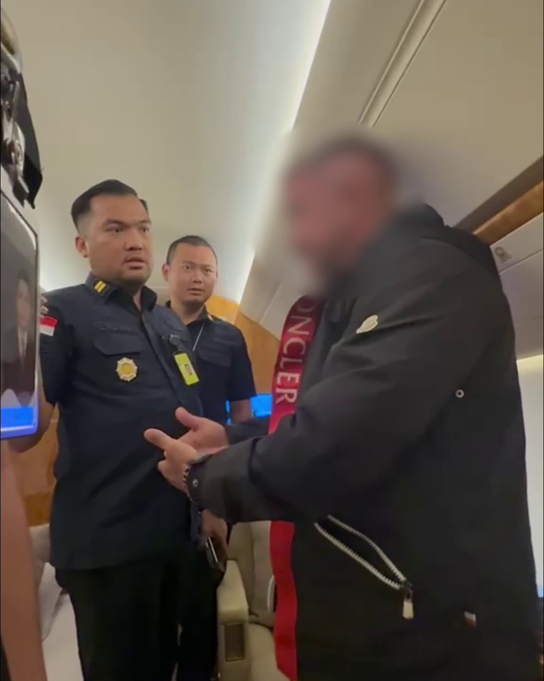 Man with face blurred speaks to immigration officers inside a private jet cabin.