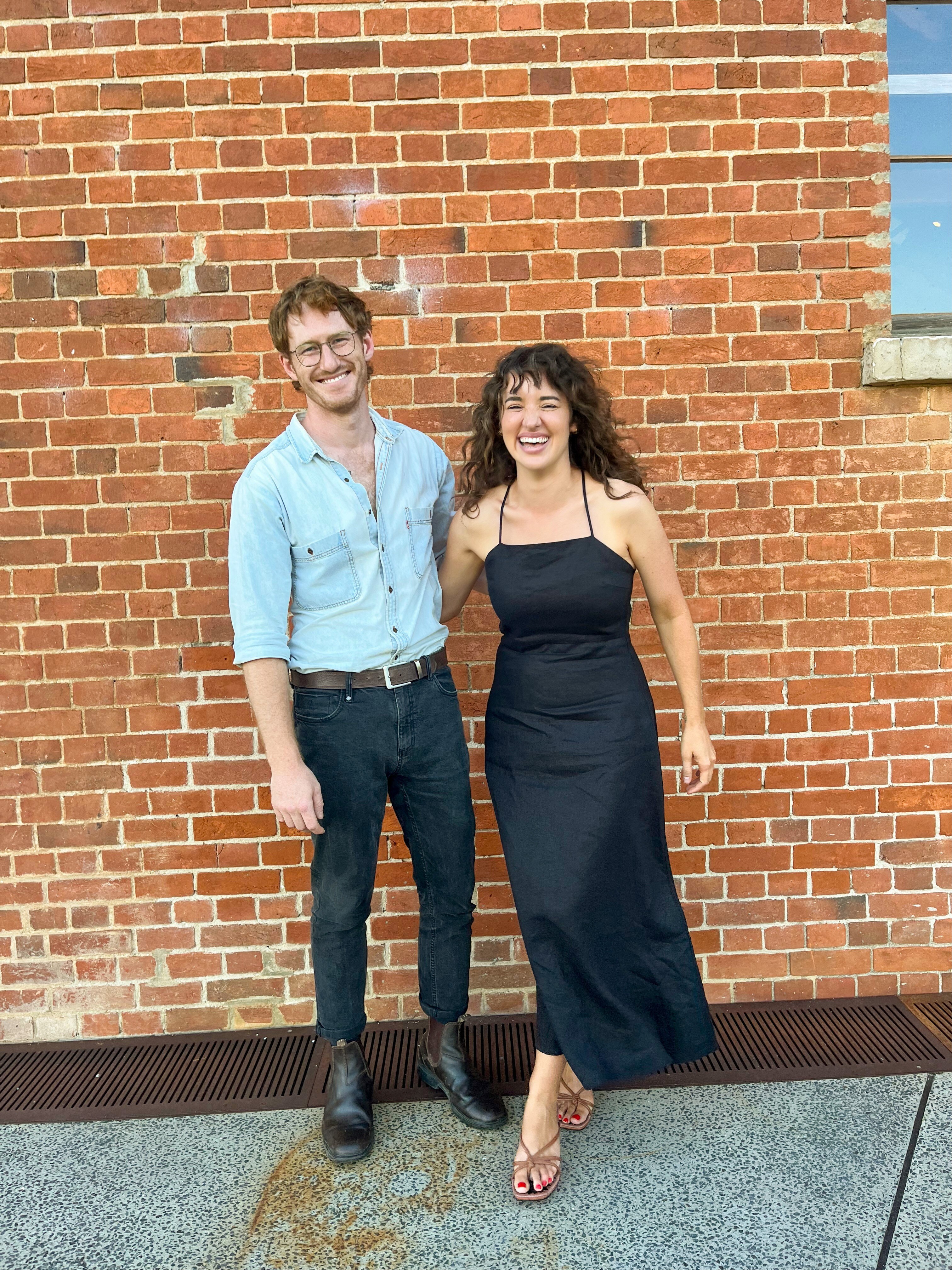 A young man and woman in formal clothes standing in front of a brick wall and smiling