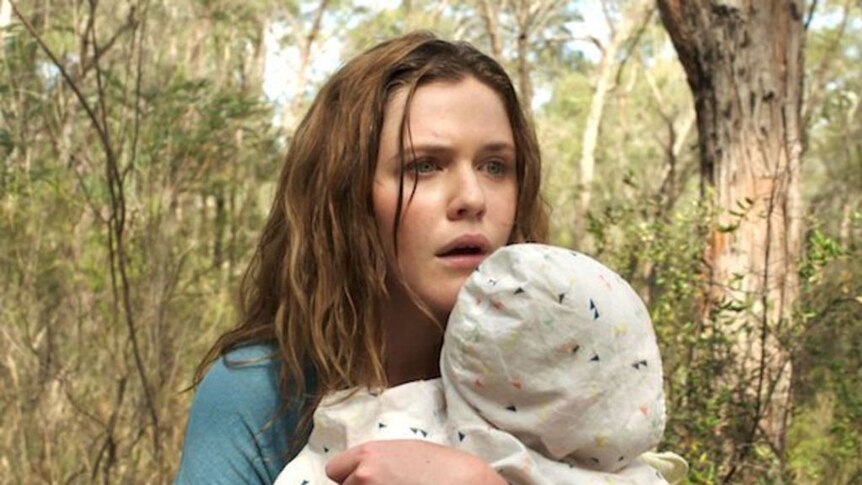Actor Harriet Dyer holds a baby while looking scared in a bush setting.