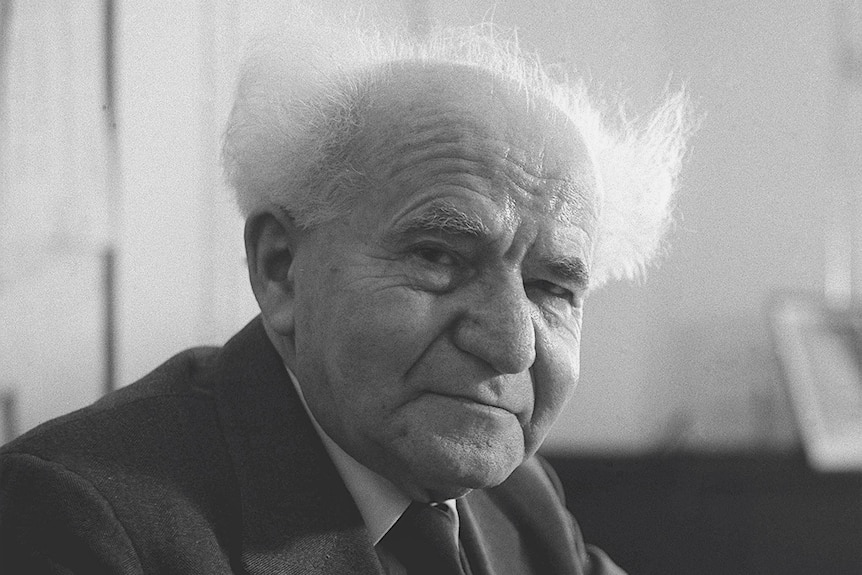 A 1960 black-and-white photo of an older man in a suit, mostly bald, but with tufts of white hair at the back