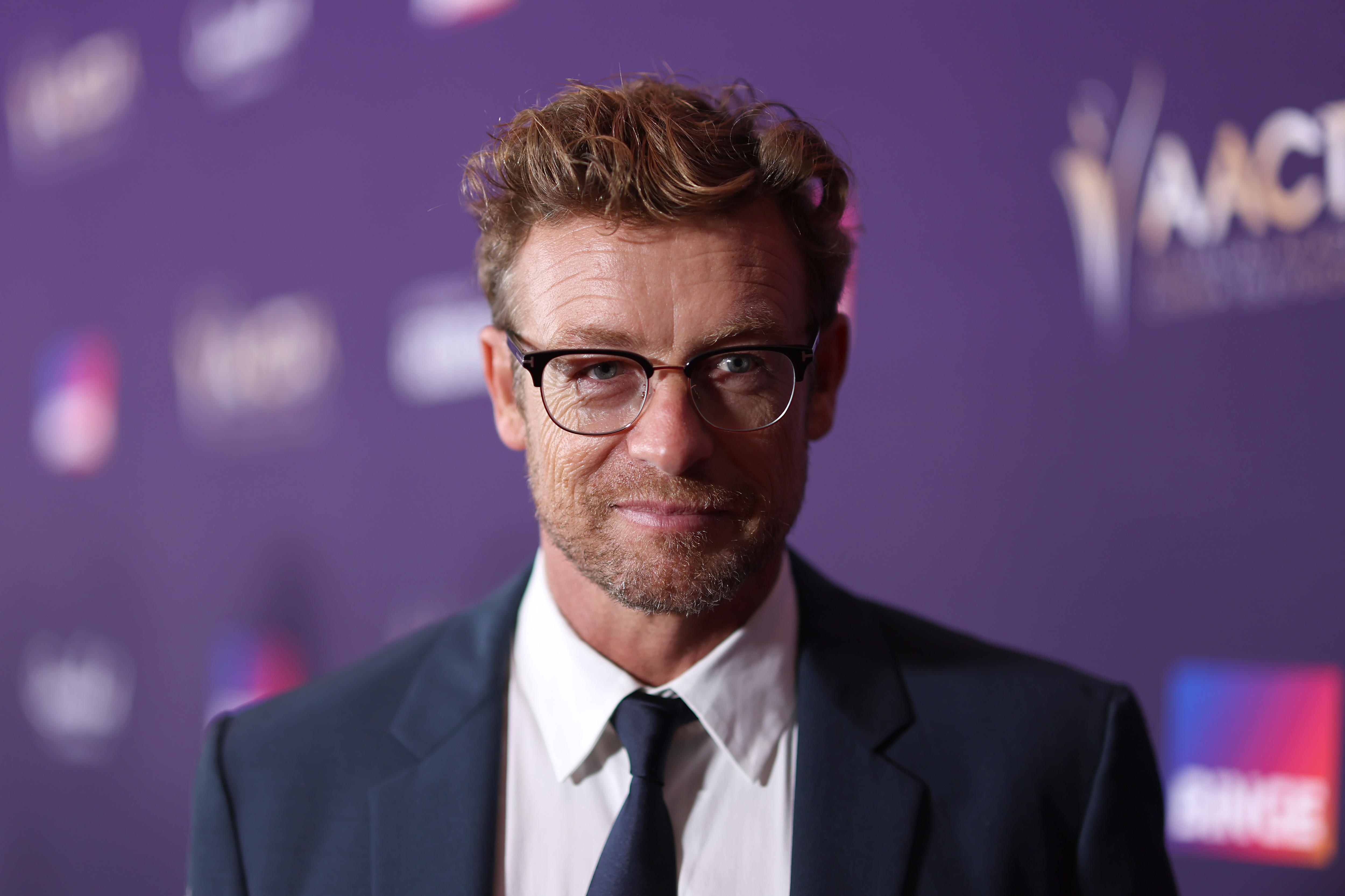 Shoulders-up picture of Simon Baker wearing a navy suit, white shirt and glasses