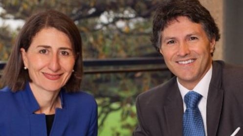 A woman in a blue jacket and a man in a suit and blue tie poise pens over documents and smile.