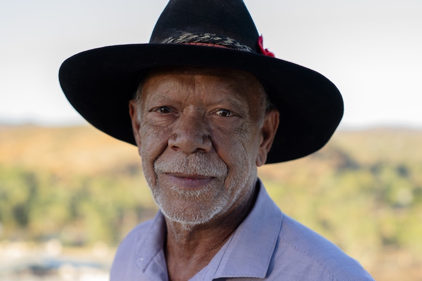 An elderly Indigenous man wearing a cowboy hat and purple shirt.