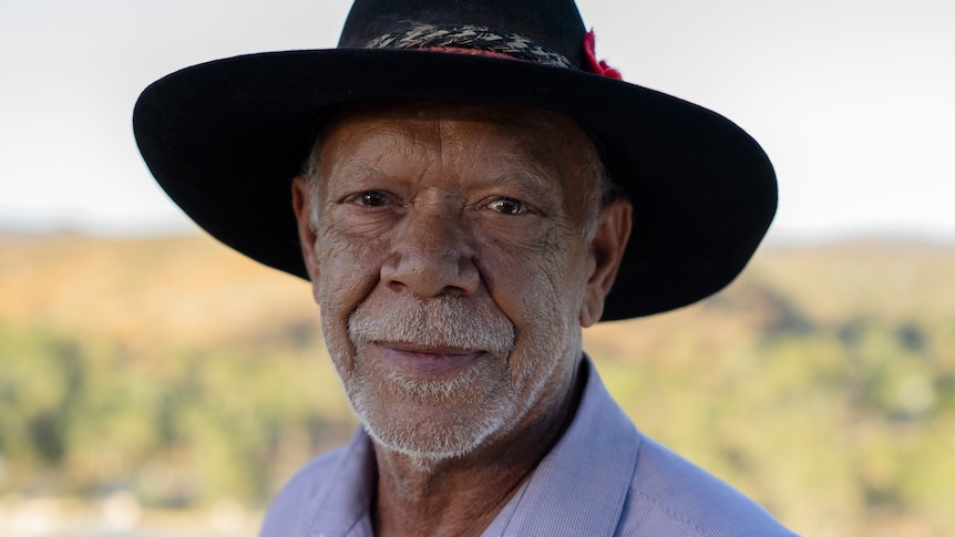 An elderly Indigenous man wearing a cowboy hat and purple shirt.
