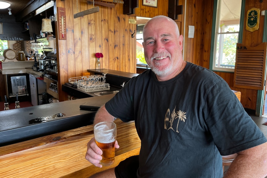 A bald man with white facial hair is smiling, holding a glass of beer and sitting at a bar at a pub.