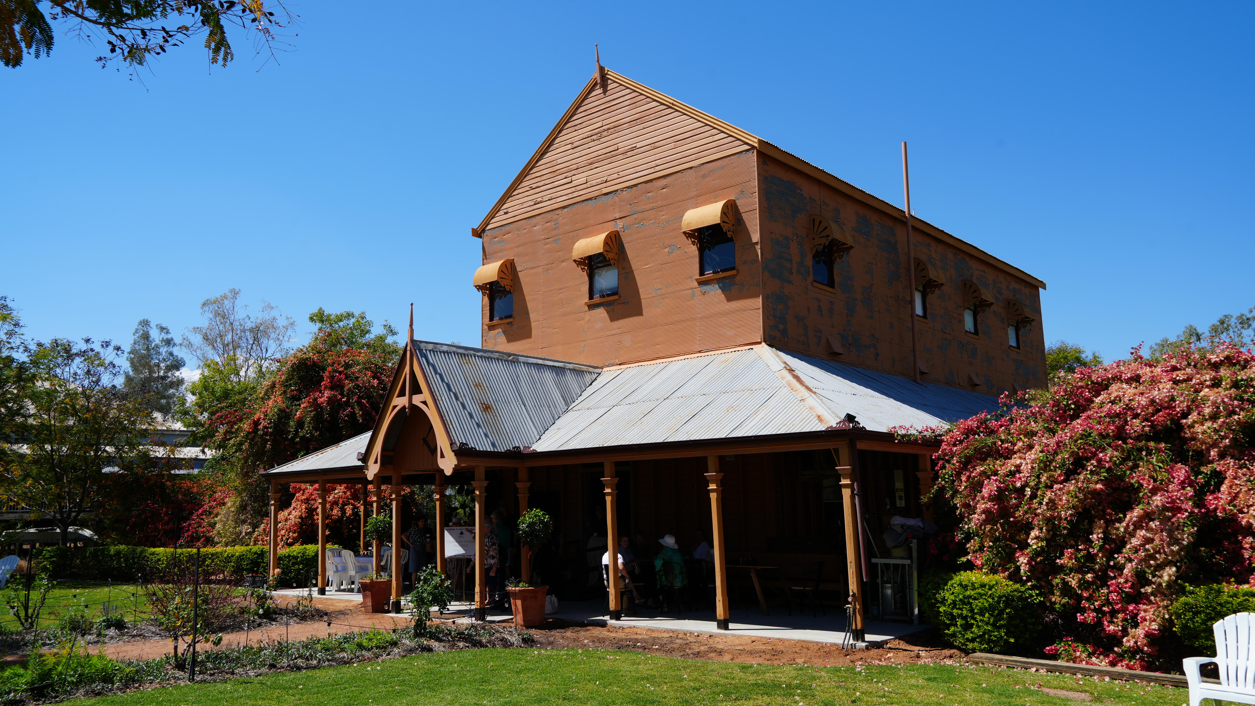 A brown tin-roofed masonic lodge surrounded by lawns and brightly coloured bushes on a sunny day.