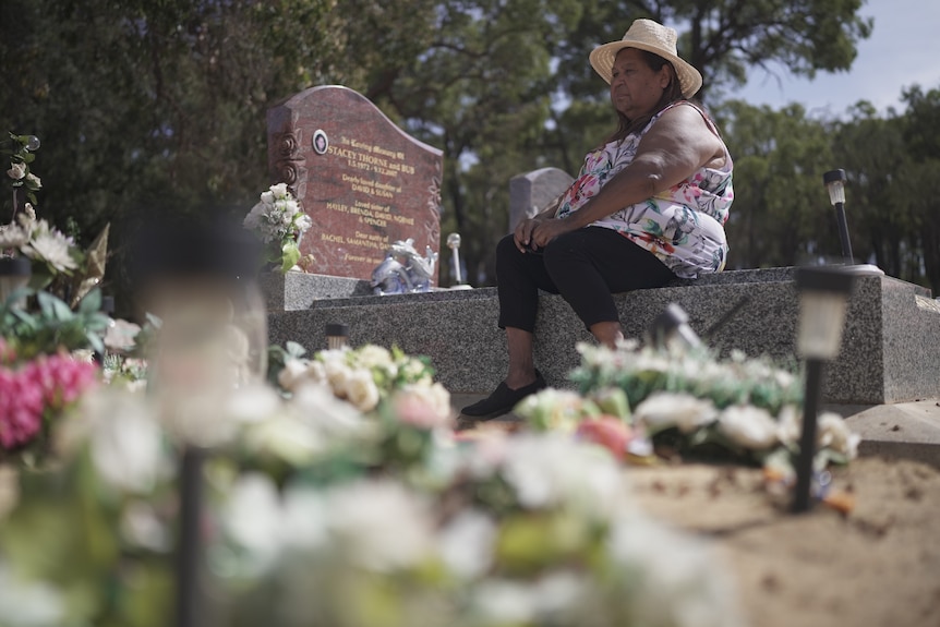 A woman in a widebrim hat sits on the edge of her sister's grave with flowers in foreground