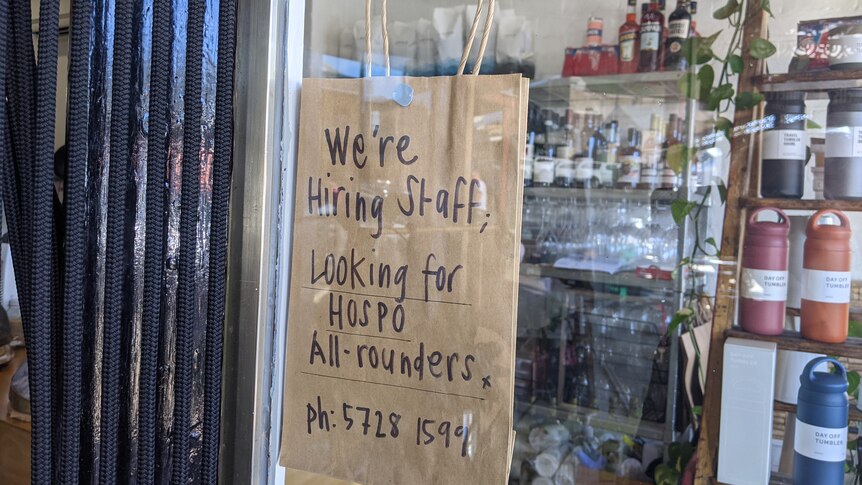 A sign on brown paper in a cafe window reads: We're hiring staff, looking for hospo all rounders