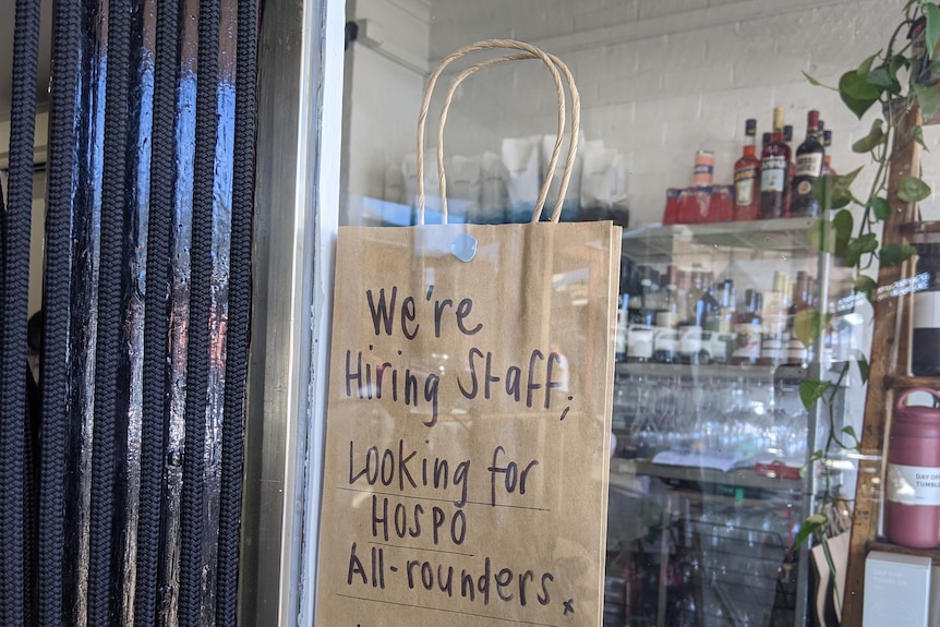A sign on a brown paper in a cafe window reads: We're hiring staff, looking for hospo' all rounders