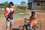 A guy stands next to a young indigenous kid on a bike