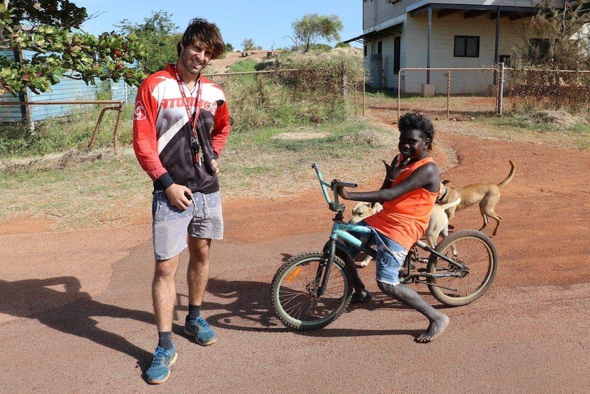 A guy stands next to a young indigenous kid on a bike