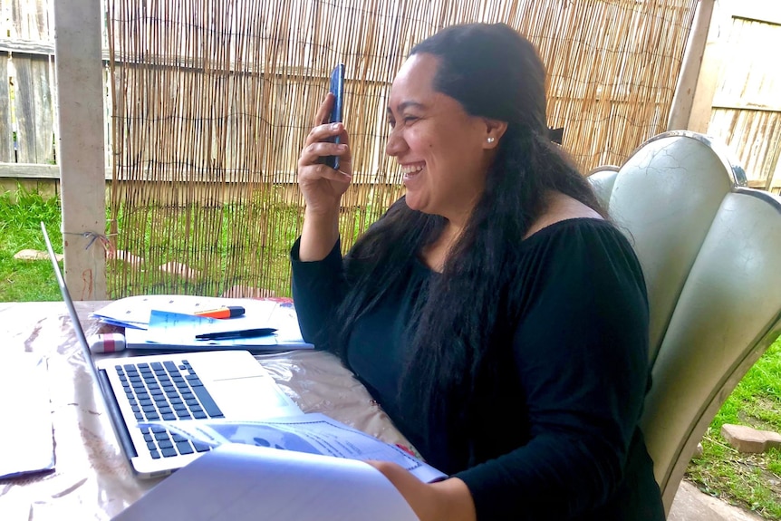 Woman on phone laughing in front of laptop, holds up papers in left hand.