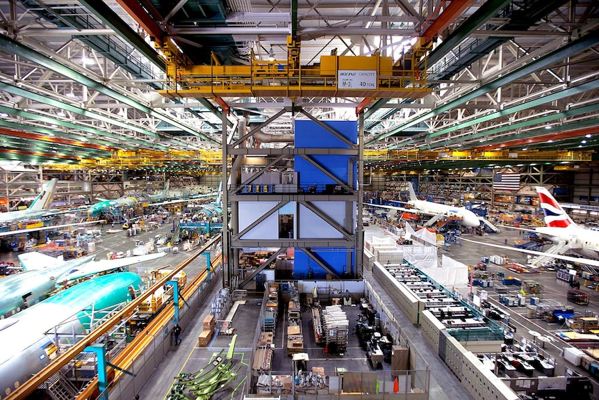 A vast wide-angle photo shows a large air hangar looking down on to a factory floor with multiple large Boeing jets.