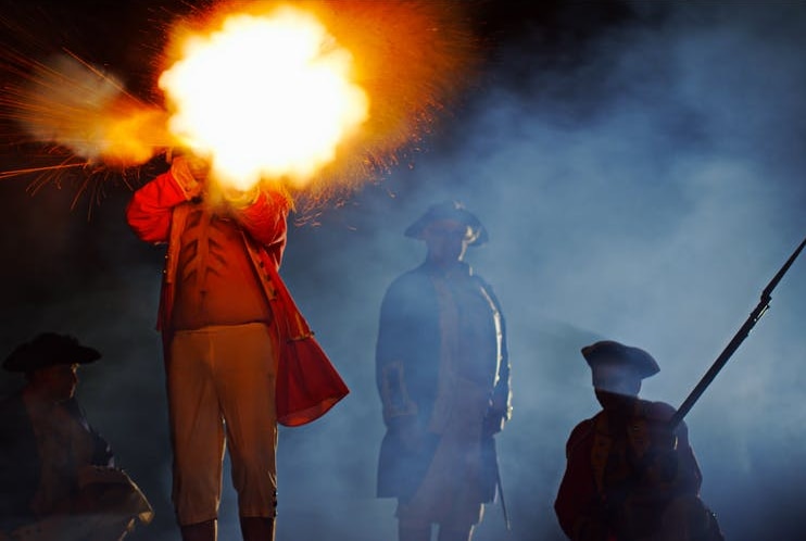 A blast of orange fire from a gun as men in historical costumes stand against a night sky.