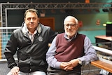 An old man with glasses and a younger man in a black leather jacket lean on a bar.