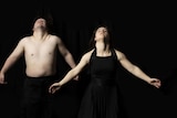 A man without a shirt and a woman in a black dress throw their heads back and arms out on a black background