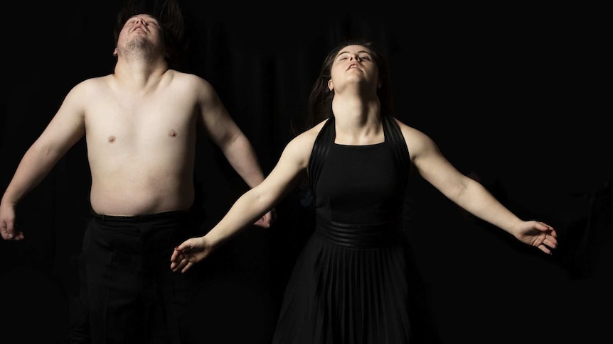 A man without a shirt and a woman in a black dress throw their heads back and arms out on a black background