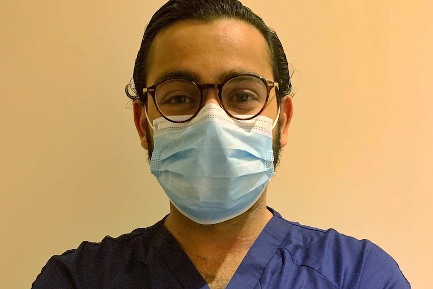 A male doctor wears dark purple scrubs and a face mask and glasses.