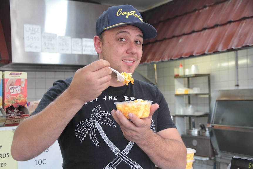 A man holds a tub of cheese slaw in one hand, a fork in the other, stands poised to take a mouthful.