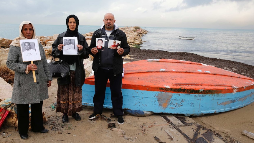 Two Muslim women and one man stand in front of an upturned boat holding pictures of their missing children