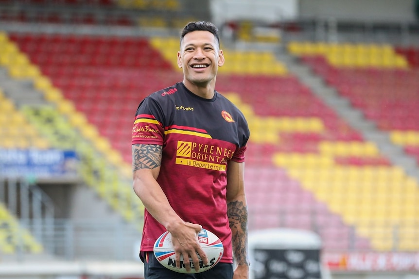 Israel Folau wears a red and black uniform on a field and stares off camera with a smile on his face.